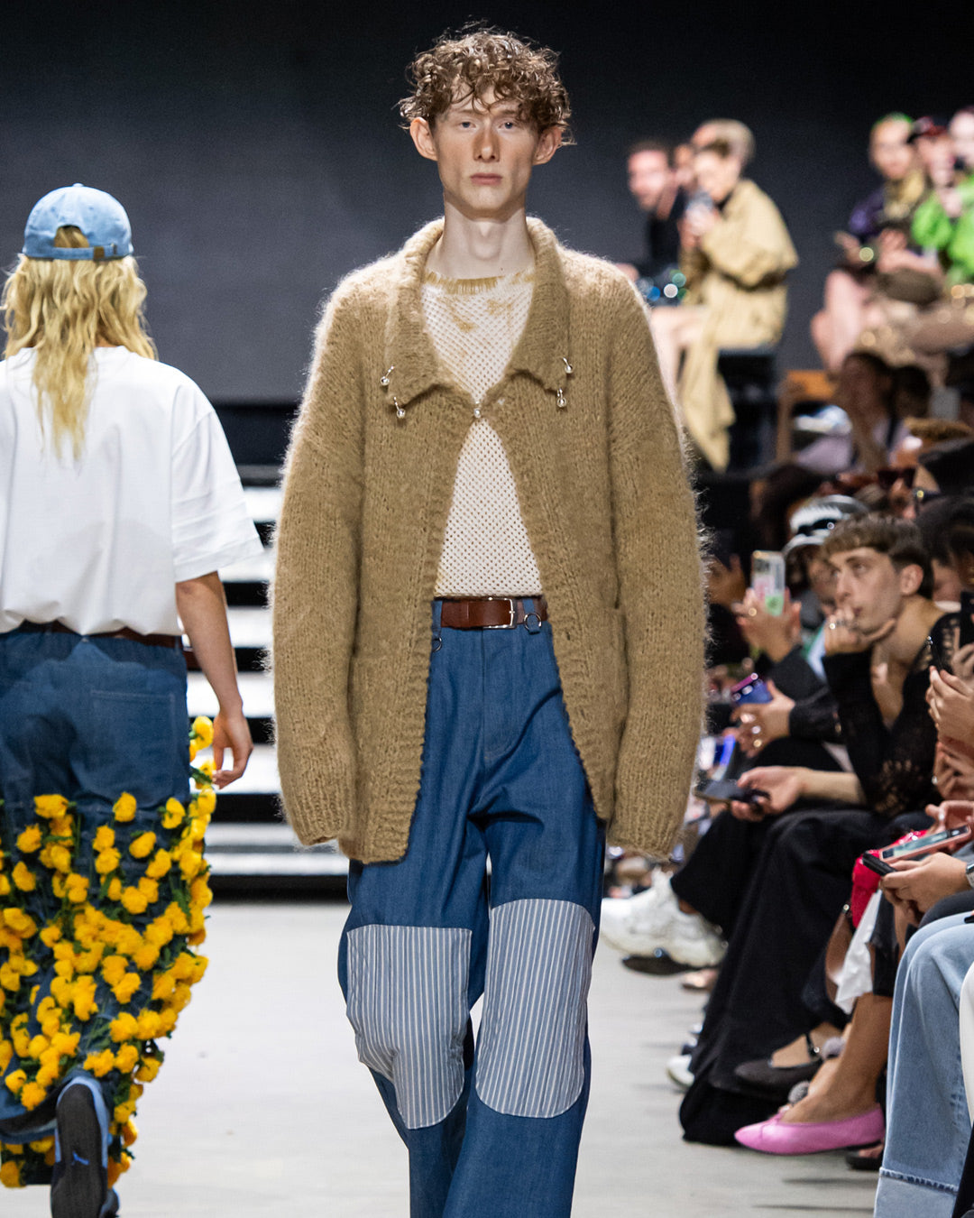 Kiharahiuksinen nuori kävelee runwayllä päällään vaaleanruskea villatakki, jossa on Rolf Ekroth x Kalevala Rakas-koruja.